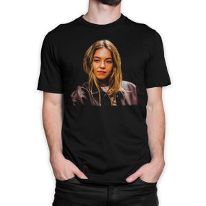 May include: Black t-shirt featuring a portrait of a woman with blonde hair wearing a brown leather jacket. The image is centered on the front of the shirt, showcasing the detailed photograph. The shirt is a classic crew neck style.