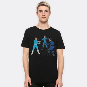 May include: Black t-shirt with a cartoon illustration of three figures in blue superhero-style outfits. One figure points, while the other two appear to be interacting. The design is outlined in black.