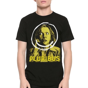 May include: Black t-shirt featuring a yellow and white graphic of a woman's face inside a smiley face. The word "happiness" is above the face, and "contagious" is below. The word "PLURIBUS" is printed in yellow.
