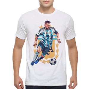 May include: White t-shirt with a graphic of a soccer player in a blue and white jersey with the number 10 on it. The player is dribbling a soccer ball and is surrounded by a gold crest with the letters "AFA".