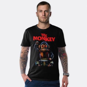May include: Black t-shirt featuring the text "THE MONKEY" in red. The graphic depicts a vintage toy monkey with red eyes, holding a knife. The monkey is wearing a red vest and sitting on a drum.