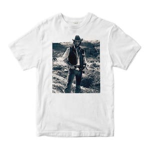 May include: White t-shirt featuring a black and white image of a man in a cowboy hat and vest standing in a desert landscape. The image is centered on the shirt, showcasing the western theme.