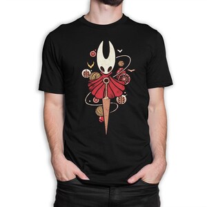May include: Black t-shirt featuring a stylized illustration of a character with a white head, red cape, and a sword. The design includes decorative elements like yarn balls and birds. The shirt is made of a soft material.