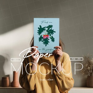 May include: A light blue paper mockup held in front of a person, featuring a botanical illustration of a plant with green leaves and a pink flower. The text "Find me in the garden" is above the illustration.