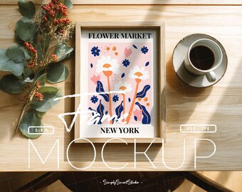 Pode incluir: Uma impressão artística emoldurada com um design floral, apresentando flores brancas e laranja, folhas azuis e o texto "Flower Market New York". A impressão está sobre uma mesa de madeira, ao lado de uma xícara de café e um galho de bagas vermelhas.