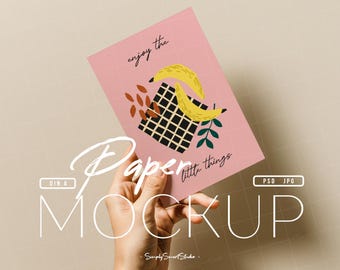 Op de afbeelding: Een roze papieren mockup in de hand, met een ontwerp van gele bananen, een zwart-wit geruit vierkant en de tekst "enjoy the little things". De mockup bevat ook de tekst "Paper Mockup".
