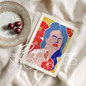 May include: A mock-up featuring a colorful illustration of a woman with blue hair, red lips, and floral accents. The artwork is on a white card, with the word "Paper" and "MOCKUP" overlaid. Cherries and a decorative plate are also in the image.