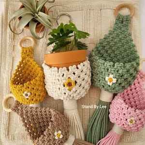 May include: Five macrame plant hangers in mustard yellow, cream, sage green, brown, and pink, each featuring a wooden ring at the top, a small flower motif, and a long tassel. The cream hanger holds a terracotta pot with a plant. Text reads Stand By Lee.
