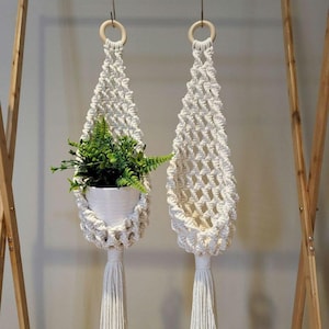 May include: Two white macrame plant hangers with wooden rings at the top and tassels at the bottom. One hanger has a small green plant in a white pot.