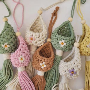 Peut inclure: Sept suspensions macramées pour plantes avec des glands et des motifs floraux de différentes couleurs. Chaque suspension a un petit panier tissé en haut.