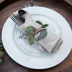 May include: A white dinner plate with a decorative pattern sits on a wooden tray. A linen napkin is wrapped around a fork and knife, and a sprig of greenery is tucked into the napkin.