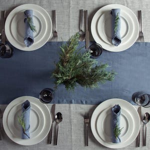 May include: A table setting with four place settings. Each setting includes a white plate, a blue napkin with a sprig of greenery, silverware, and a dark glass. A blue runner and a green plant are in the center.