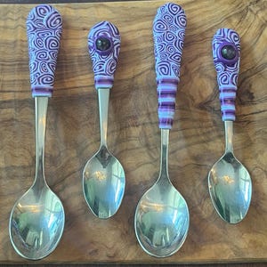 May include: Four silver spoons with purple and white swirl patterned handles. The spoons are arranged on a wooden surface.