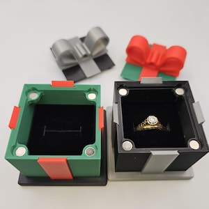May include: Two decorative ring boxes, one green and one black, with red accents and magnetic closures. The black box holds a gold ring with a diamond. Two gift box-shaped toppers with bows are in the background.