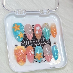 May include: A set of ten press-on nails with a sea-themed design. The nails feature various colors, including blue, pink, and orange, with embellishments like starfish, pearls, and seashell designs. The nails are in a clear plastic case.