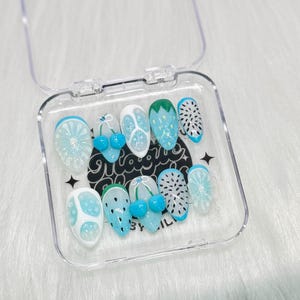 May include: A set of ten blue and white press-on nails with fruit designs. The nails feature watermelon, cherries, and strawberries. The nails are in a clear plastic container with the text "Magic By Nail" on the bottom.