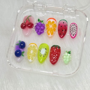 May include: A clear plastic container with 10 colorful fruit-shaped nail decorations. The decorations include cherries, grapes, kiwi, strawberries, watermelon, and lemons.