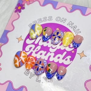 May include: A set of press-on nails with colorful fruit designs, including lemons and oranges. The nails feature a variety of patterns and colors, such as orange and white stripes, and blue and white floral designs. The nails are displayed on a decorative card.
