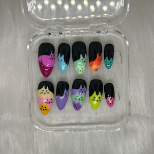 May include: A set of press-on nails in a clear plastic case. Each nail is painted black at the top and features a colorful cat face design. The colors include pink, blue, green, orange, and yellow.