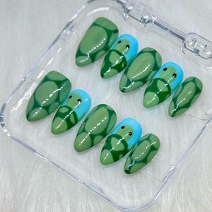 May include: A set of ten green and blue false nails with a turtle design. The nails are in a clear plastic case.