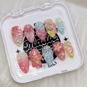 May include: A set of ten artificial nails in a white case. The nails feature various designs, including floral patterns, gradients, and embellishments like pearls and small flowers. The nails are in shades of pink, blue, yellow, and white.