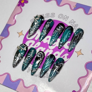 May include: A set of ten long, pointed press-on nails. Each nail features a teal and black gradient design with silver accents and pearl embellishments. Some nails have intricate silver filigree designs. The nails are arranged on a decorative card.
