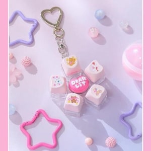 Puede incluir: Un llavero con un botón circular central de color rosa brillante con el texto DEAR LIT en blanco, rodeado por cinco teclas cuadradas de color rosa claro decoradas con ilustraciones de osos y fresas, unido a un cierre plateado en forma de corazón.