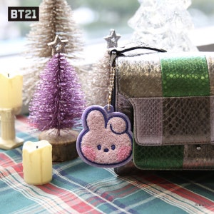 May include: A pink and purple bunny-shaped keychain with a fluffy texture hangs from a silver chain attached to a multi-colored purse. The purse is made of a patchwork of different materials, including snakeskin and leather. The background features a purple and silver Christmas tree.