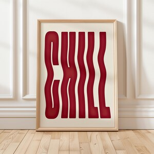 May include: A framed art print with the word "CHILL" in a wavy, bold, red font against a cream background. The wooden frame adds a natural touch. The artwork is displayed on a wooden floor against a white wall.