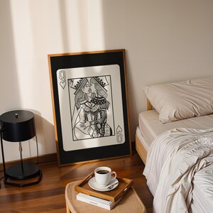 Puede incluir: Una impresi&oacute;n art&iacute;stica en blanco y negro enmarcada de dos mujeres bes&aacute;ndose, dise&ntilde;ada como una carta de juego. La impresi&oacute;n est&aacute; apoyada contra una pared en un dormitorio, con una cama, una mesita de noche y una l&aacute;mpara.