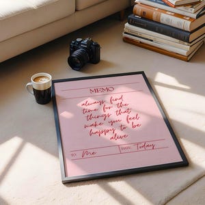 May include: A framed pink memo with the text "always find time for the things that make you feel happy to be alive." A black camera, a coffee mug, and a stack of books are also visible.