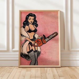 May include: Pop art style print of a woman in lingerie holding an orange chainsaw. The artwork features a pink background and is framed in a light wood frame. The woman has black hair and is wearing a black bra and garter belt.