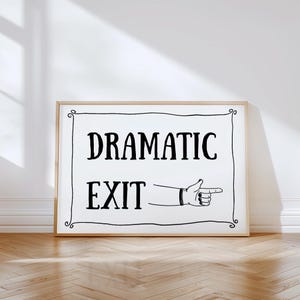 May include: A framed sign with the words "DRAMATIC EXIT" in bold black letters. Below the text is a drawing of a hand pointing to the right. The sign has a decorative border and is set against a white wall.