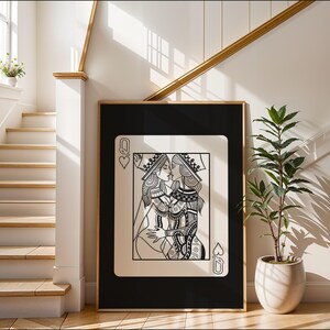 Puede incluir: Una impresi&oacute;n art&iacute;stica enmarcada que presenta una ilustraci&oacute;n en blanco y negro de dos reinas bes&aacute;ndose, sobre un fondo negro. La obra de arte est&aacute; dise&ntilde;ada como una carta de juego, con el s&iacute;mbolo de la Reina de Corazones visible. El marco es dorado y la impresi&oacute;n se exhibe en un interior moderno y luminoso.