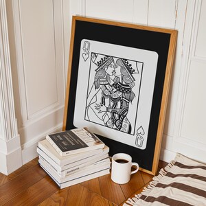 Puede incluir: Una impresi&oacute;n art&iacute;stica enmarcada que presenta una ilustraci&oacute;n en blanco y negro de dos reinas bes&aacute;ndose, al estilo de una carta de juego. La obra de arte est&aacute; enmarcada en madera, apoyada contra una pared. Una pila de libros y una taza de caf&eacute; est&aacute;n en primer plano.