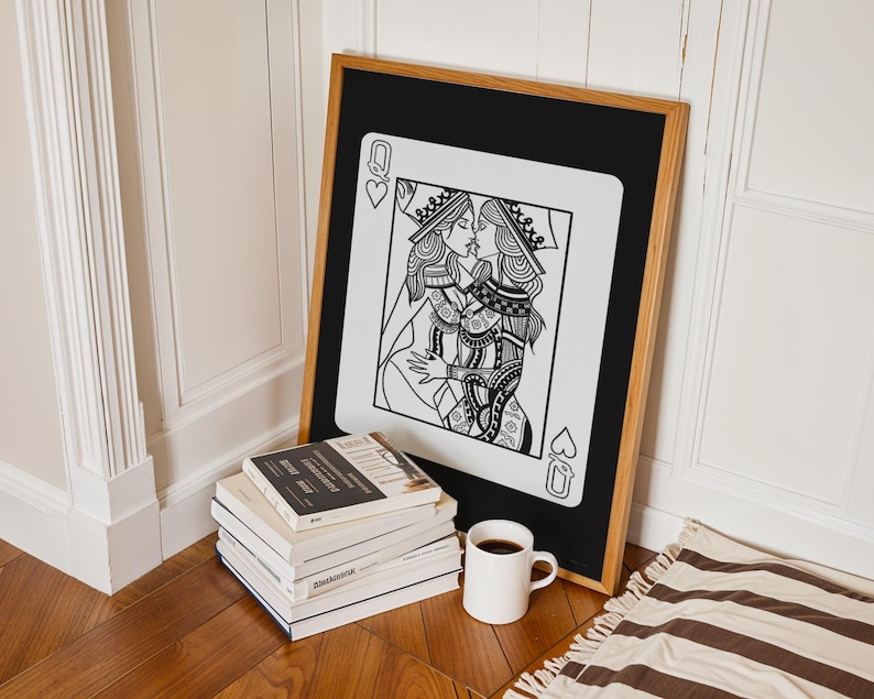 Puede incluir: Una impresi&oacute;n art&iacute;stica enmarcada que presenta una ilustraci&oacute;n en blanco y negro de dos reinas bes&aacute;ndose, al estilo de una carta de juego. La obra de arte est&aacute; enmarcada en madera, apoyada contra una pared. Una pila de libros y una taza de caf&eacute; est&aacute;n en primer plano.