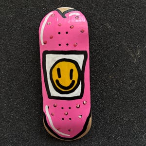 May include: A fingerboard deck featuring a bright pink base with a white rectangular patch in the center containing a yellow smiley face with black eyes and a mouth. The pink surface is decorated with small red dots and white highlights.