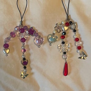 May include: Two beaded phone charms with pink, purple, red, and white beads. Each charm features heart and star-shaped beads, clear bow accents, and small butterfly charms. The charms have black loops for attachment.