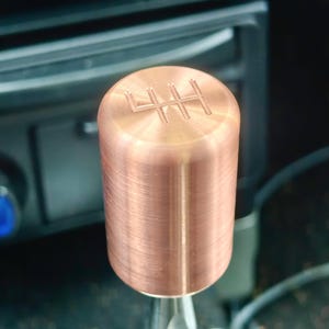 May include: A copper-colored shift knob with a 4-speed pattern engraved on the top. The cylindrical knob is mounted on a silver-colored shift lever. The background shows a car's interior.