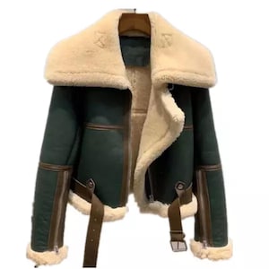 Può includere: Una giacca di pelle di pecora verde con finiture in pelle marrone e un ampio colletto in pelliccia bianca. La giacca ha una chiusura a zip e una cintura in vita.