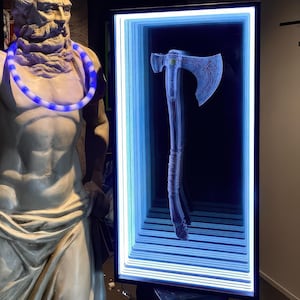 May include: A white statue of a man holding a golden trident, wearing a blue light-up necklace, standing in front of a framed artwork of a silver axe with a black handle.