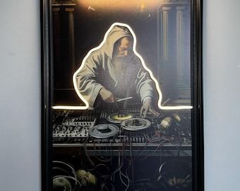 May include: Framed artwork depicting a robed figure with a long beard, illuminated by a glowing outline, operating DJ equipment. The artwork is in a black frame, and a gold-toned picture light is mounted above it.