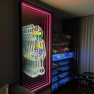 May include: A neon sign depicting a multi-coloured robot figure in a black frame with pink and white neon outlines. The sign is mounted on a wall in a room with a glass cabinet filled with toy cars.
