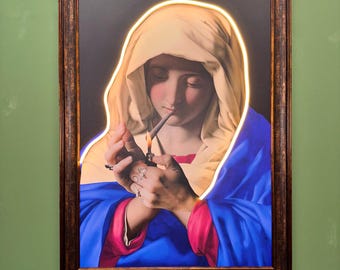 May include: Framed artwork featuring a religious figure lighting a cigarette. The figure is wearing a white head covering and blue and pink robes. A neon light outlines the figure. The frame is brown.