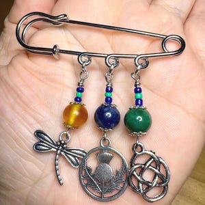 May include: A silver safety pin with three charms: a dragonfly, a thistle, and a Celtic knot. Each charm is attached to a beaded chain with a gemstone bead: amber, lapis lazuli, and green aventurine.