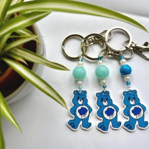 Peut inclure: Trois porte-clés en forme d'ours bleu Care Bear avec un nuage blanc et des gouttes de pluie sur le ventre. Chaque porte-clés a un anneau argenté et un charme perlé avec une perle bleue et blanche.