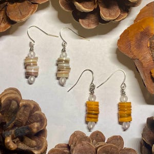 May include: A pair of earrings featuring a stack of iridescent shell beads in a light brown and white colour and a stack of yellow shell beads. The earrings are silver and have a hook fastening.
