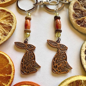 May include: Two copper rabbit keychains with intricate designs, each with a silver key ring and a beaded charm featuring orange and brown wooden beads and clear glass beads.