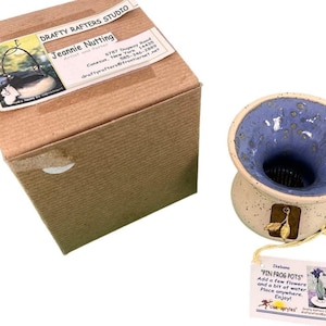 May include: A ceramic pin frog pot with a blue interior and a speckled exterior, next to a cardboard box with a label that reads "DRAFTY RAFTERS STUDIO Jeannie Nutting". A tag attached to the pot reads "PIN FROG POTS".