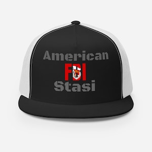 May include: A black and white trucker hat with a black bill and a white mesh back. The hat features the text "American FBI Stasi" in grey lettering with the word "FBI" in red.
